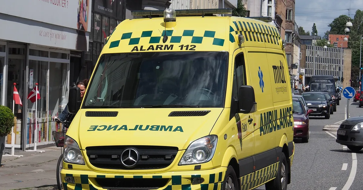 Gul dansk ambulanse merket «ALARM 112» i en bygate